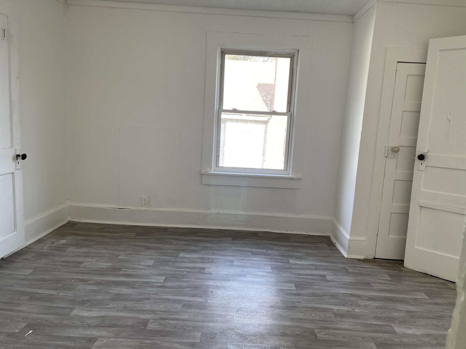 An empty room with white walls, a single window, wooden laminate flooring, and two closed white doors.