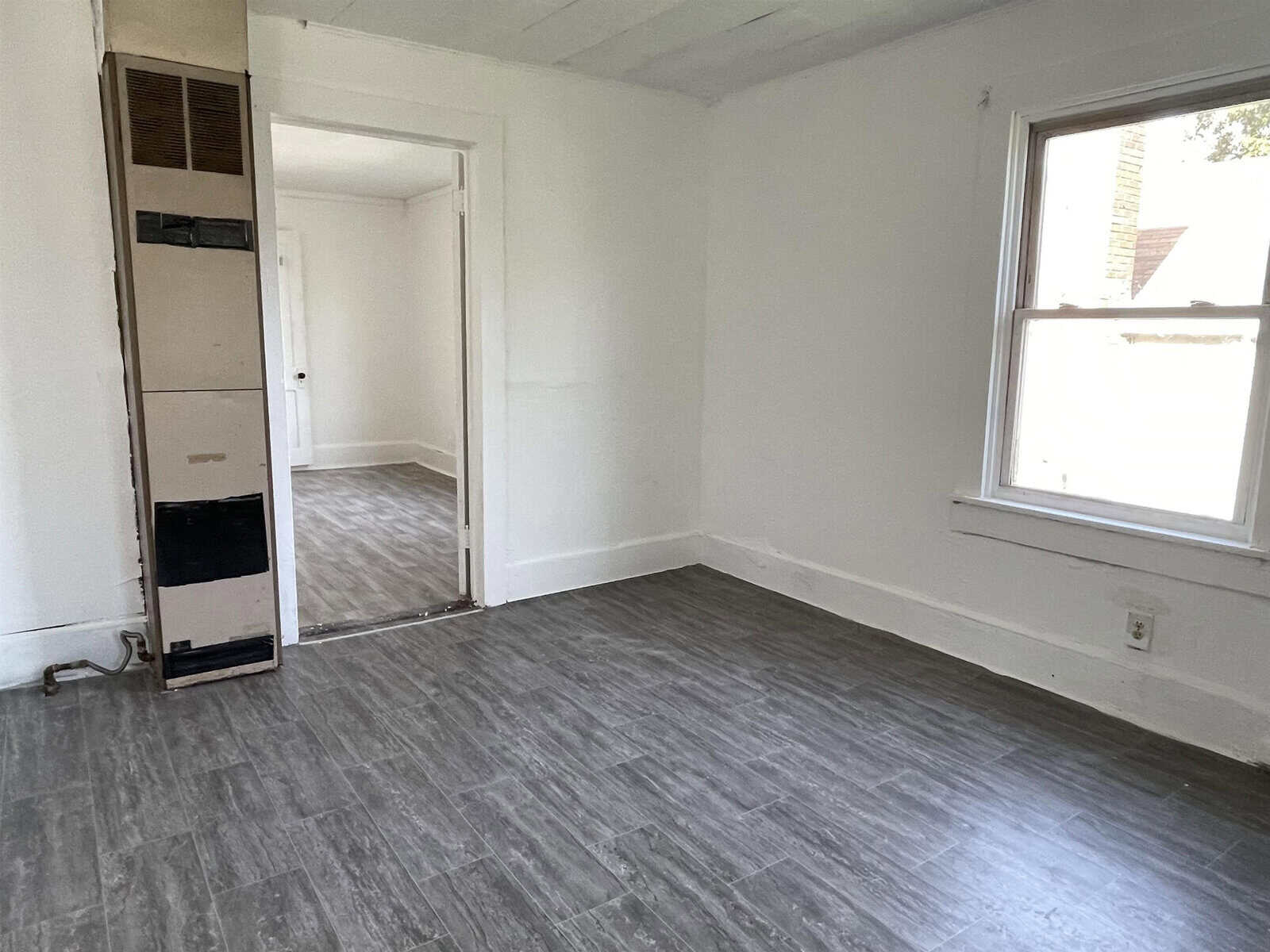 An empty room with white walls and gray flooring, featuring a doorway leading to another room and a window allowing natural light.