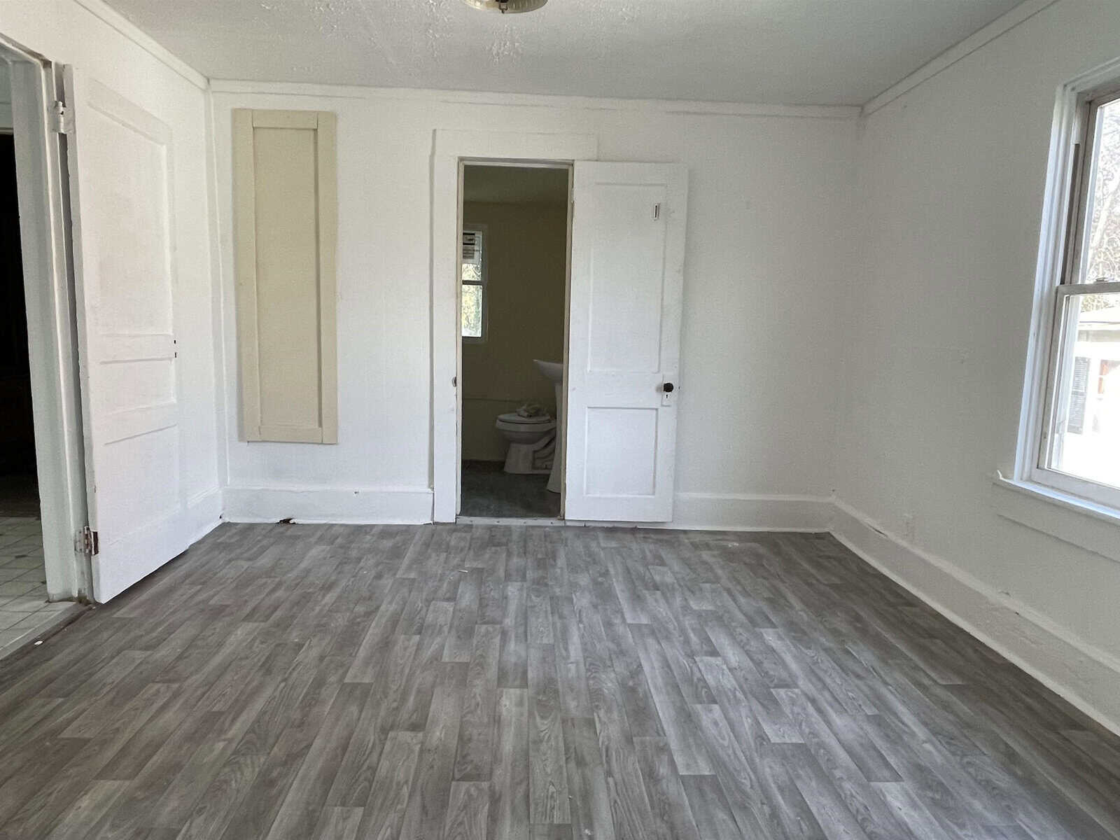 An empty room with gray flooring, white walls, and two closed doors, one of which is ajar and leads to a bathroom with a visible toilet.