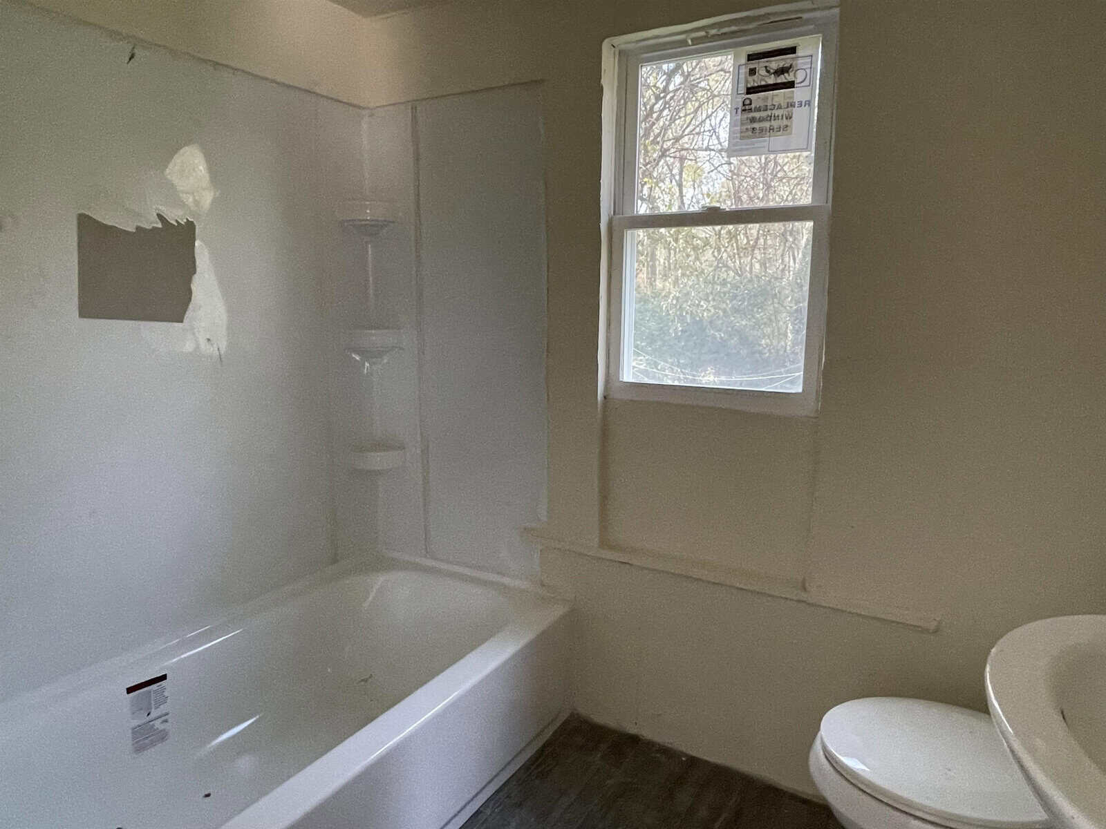 A bathroom in need of maintenance, featuring a tub, toilet, and a window with a visible sticker, with some wall damage and signs of wear.
