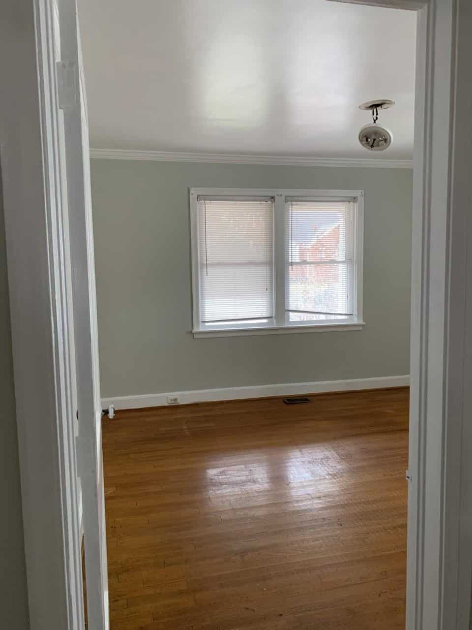 An empty room with light green walls, hardwood floors, a pair of windows with blinds, and a ceiling light fixture, viewed from a doorway.