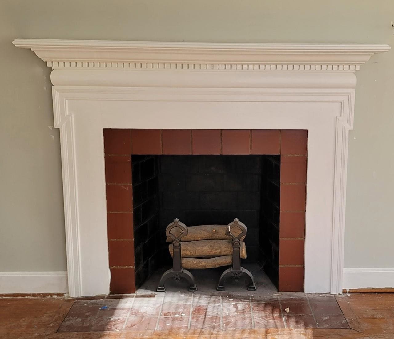A traditional fireplace with a white mantel and red brick surround, featuring an iron log holder inside the firebox, set against a green wall in a room with hardwood floors.