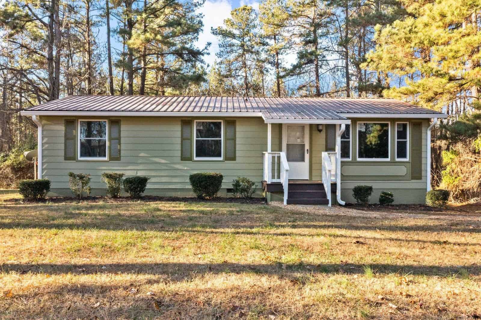 A single-story house with light green siding, a gray shingled roof, and white trim, featuring a small porch with stairs and surrounded by a few trimmed bushes and pine trees.