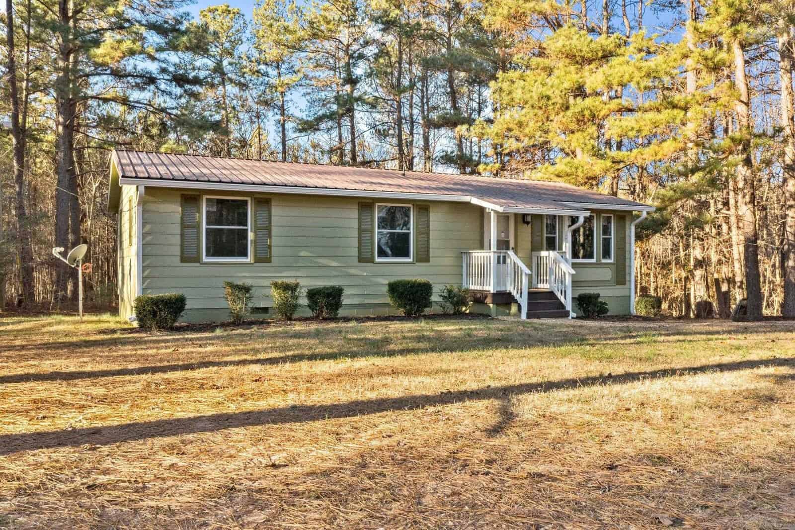 A single-story green house with white trim, featuring a small front porch with stairs, sits surrounded by a yard and trees under a clear sky.