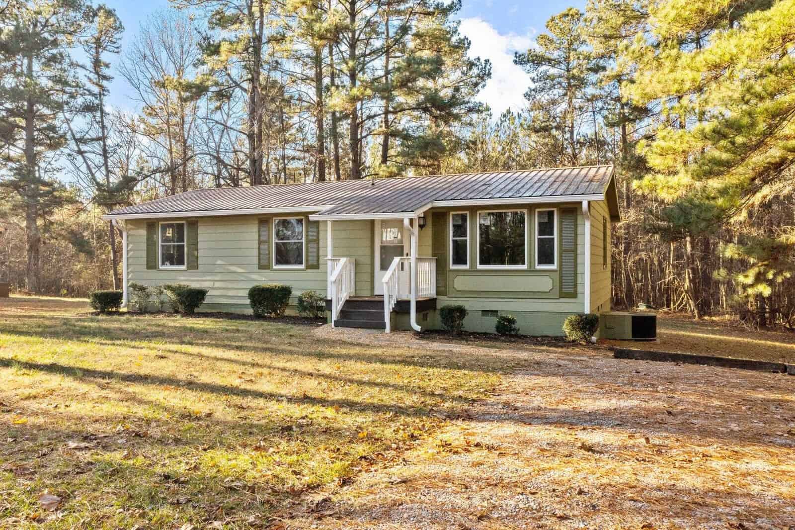A single-story house with a light green exterior, white trim, and a gable roof, situated in a wooded area with a front yard lacking grass coverage.