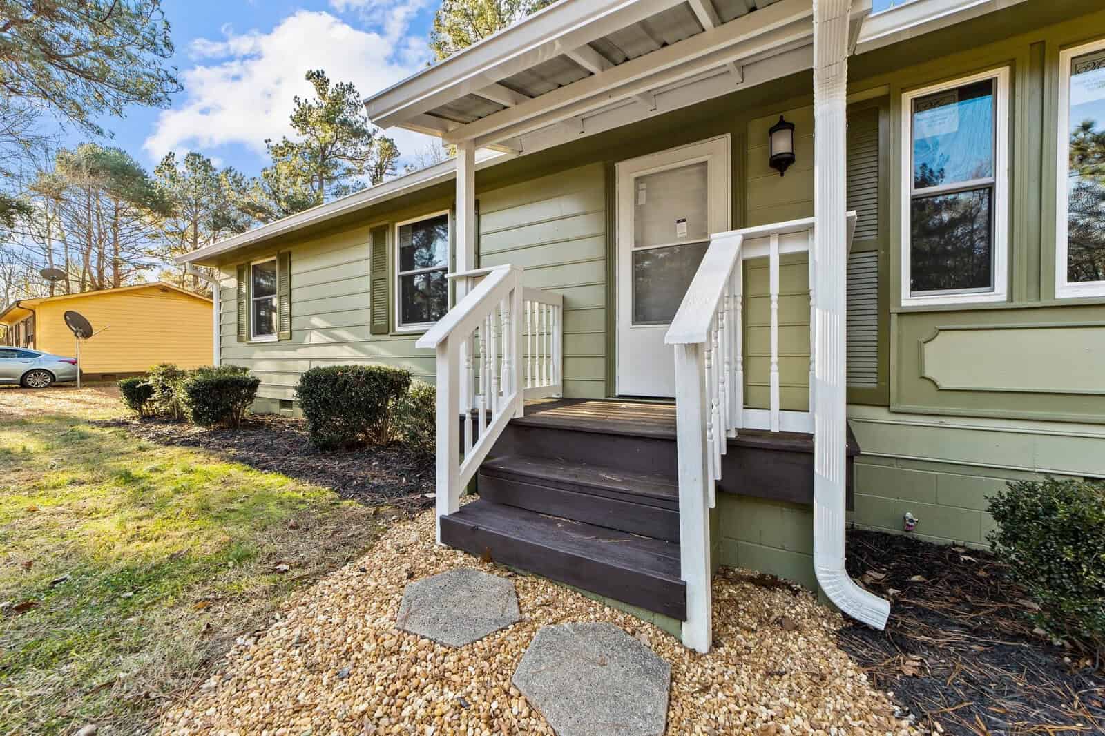 The image shows the exterior of a single-story house with olive green siding and white trim, featuring a small porch with steps leading up to a white front door. to the right, there are two large windows with visible reflections, and to the left, the yard extends with scattered trees and a shed in the distance. the roof is visibly covered in shingles, and there appears to be a driveway on the left with a parked car. the landscaping includes a bed of mulch at the base of the house front with stepping stones leading away from the porch.