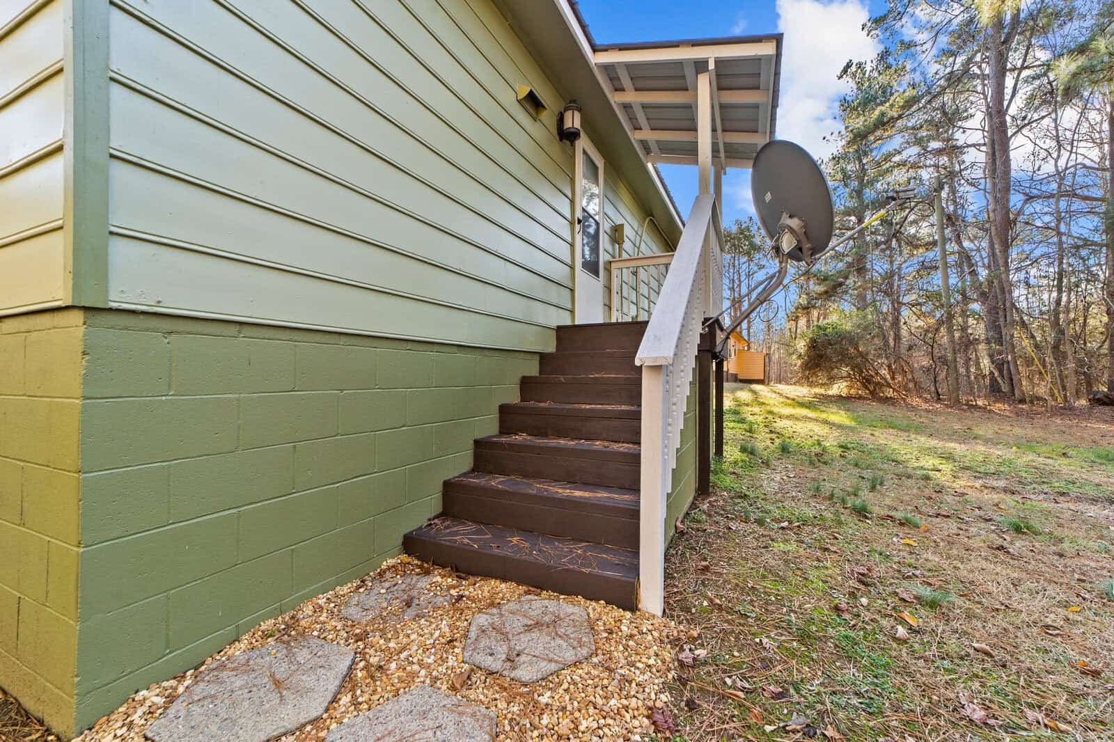 A side view of a green house with brown steps leading to a back door, a satellite dish attached near the roof, and a partly wooded area in the background.