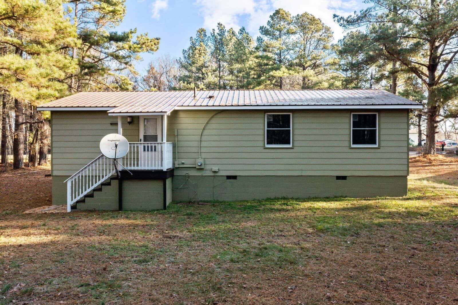 A single-story house with olive green siding and a metal roof, featuring two windows and a white door accessible by a small staircase with a railing. there's a satellite dish mounted on the side, and the house is surrounded by a grassy area with pine trees in the background.