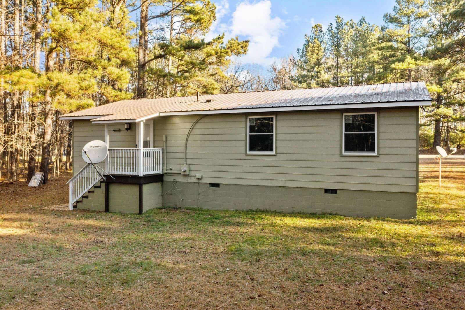 A single-story house with beige siding and a black roof, featuring a satellite dish on the side, is surrounded by a grassy yard with trees in the background on a sunny day.