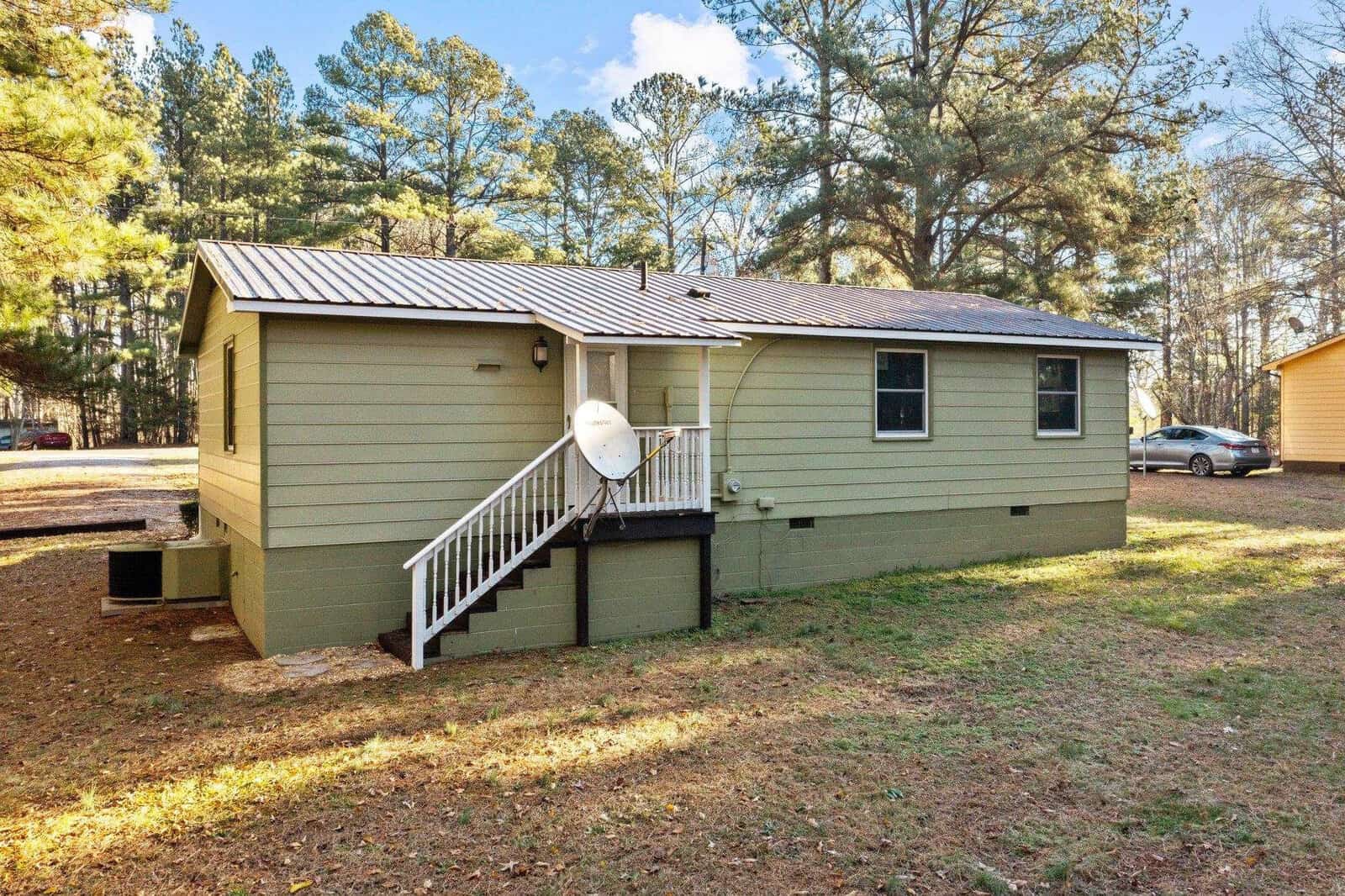 A single-story green house with white trim, featuring a satellite dish, a set of stairs leading to a back entrance, and surrounded by tall pine trees under a clear sky. a sedan car is parked to the left side.