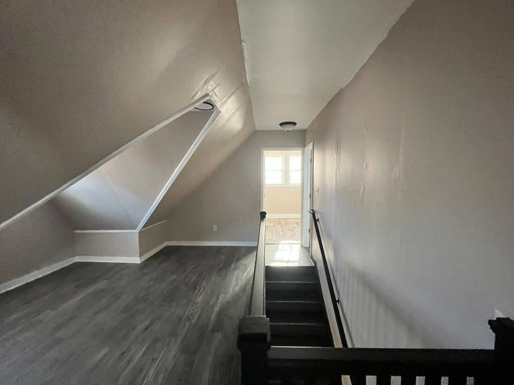 A narrow staircase with dark steps leading down to a room with natural light coming through a window, flanked by white walls and a slanted ceiling.