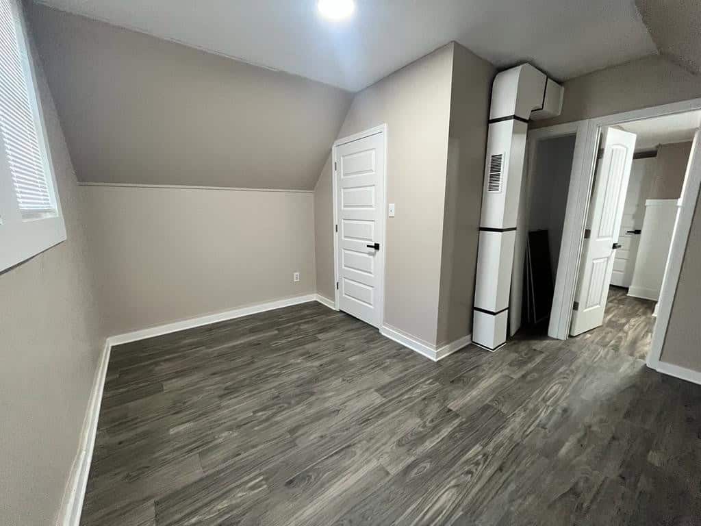 An empty room with gray laminate flooring featuring a white refrigerator beside a closed white door, with another open door leading to an adjacent room visible in the background.