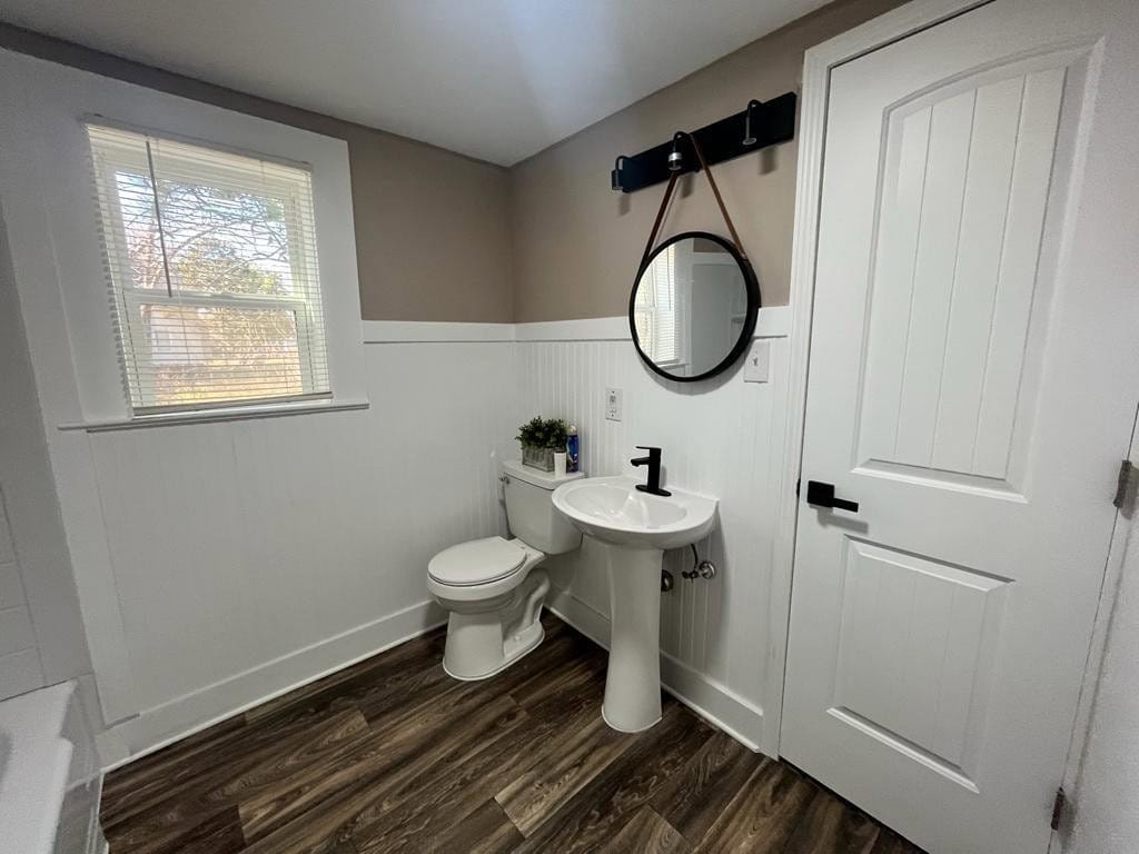 A modern bathroom featuring a white toilet and pedestal sink, with a round mirror mounted on the wall, a dark towel holder, white paneling, and wood-effect flooring.
