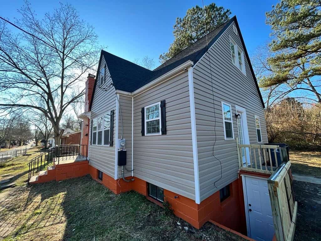 A two-story residential house with beige siding and a red foundation, featuring a small wooden deck and surrounded by a fence, under a clear sky.