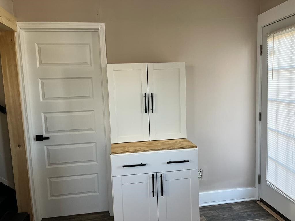 A white storage cabinet with a wooden countertop is positioned in the corner of a room next to a closed white door, with a window on the adjacent wall.