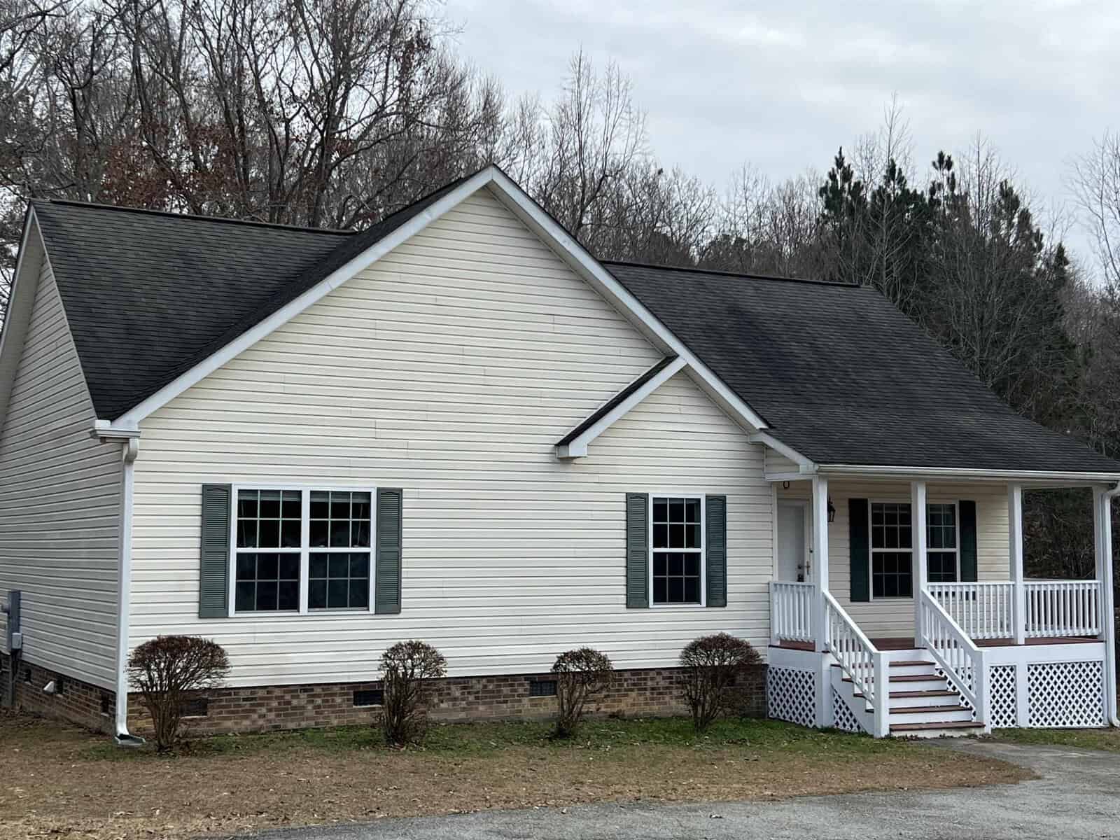A single-story white siding house with a gabled roof, featuring a small front porch with white railings, and two visible windows. the house is surrounded by bare shrubs and a partly cloudy sky overhead.