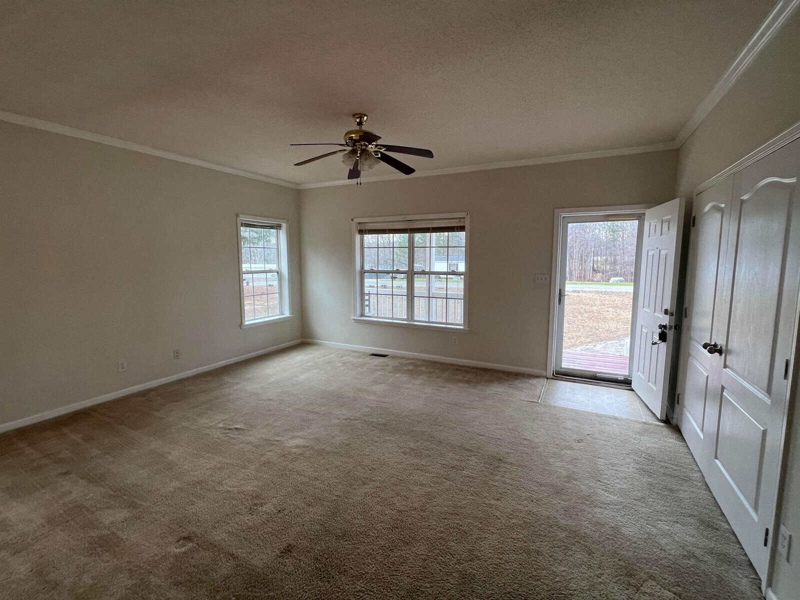 An empty room with beige carpeting, light-colored walls, a ceiling fan, and a set of windows overlooking the outside.