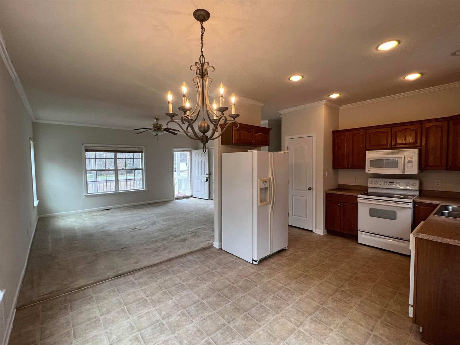 A spacious kitchen with wooden cabinets and vinyl flooring extends into a living area with carpet and a chandelier.