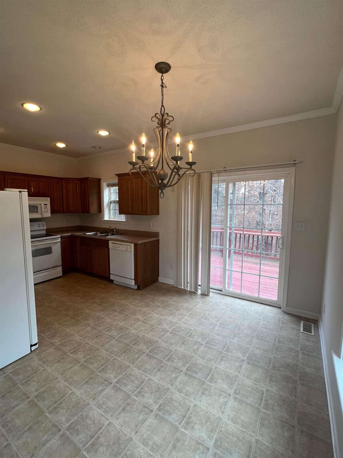 An empty kitchen with wooden cabinets, white appliances, and a tiled floor leading to a sliding glass door that opens onto a wooden deck. a chandelier hangs from the ceiling in the dining area.