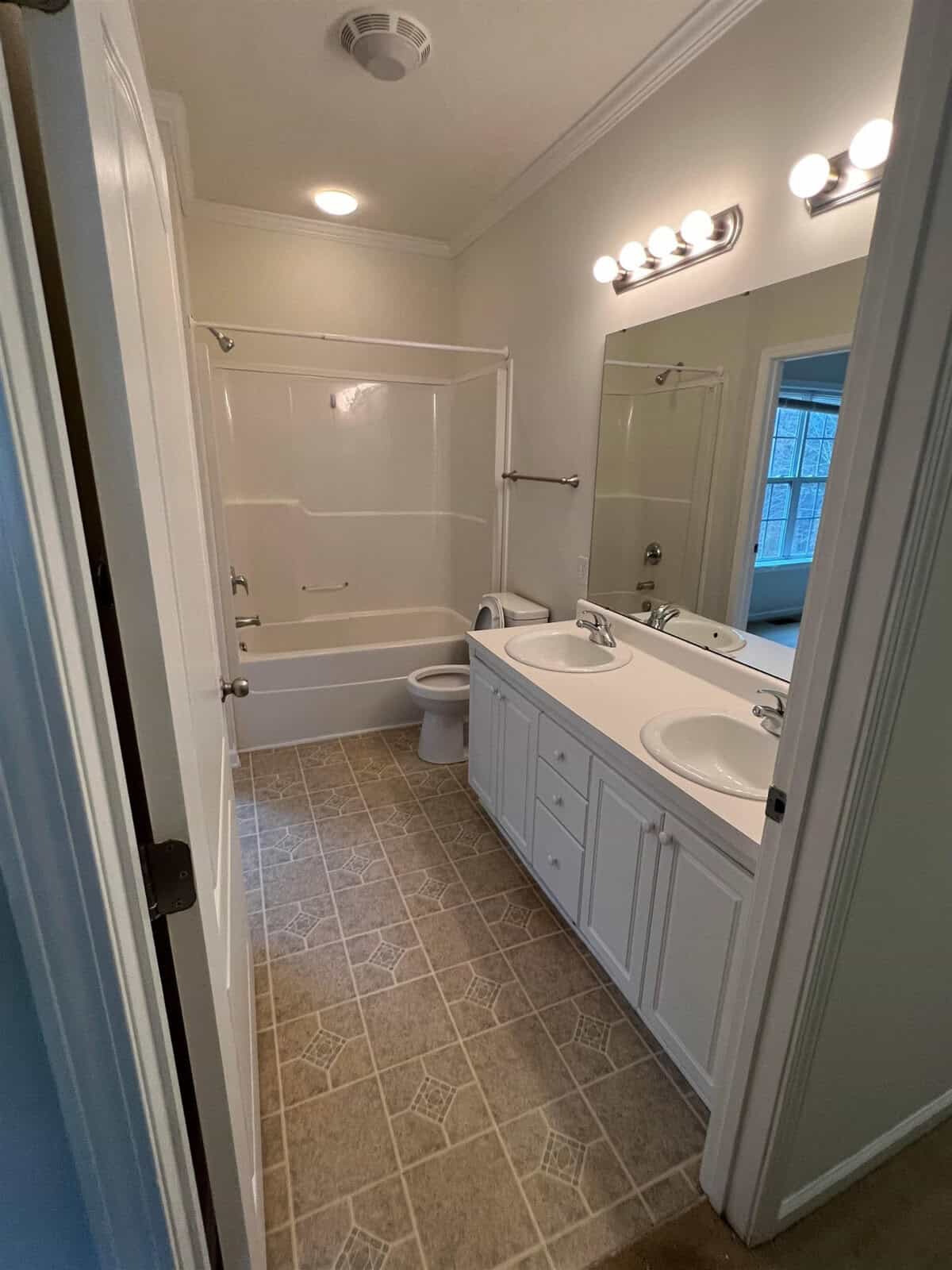 A clean, well-lit bathroom with a dual sink vanity, a large mirror with overhead lights, a toilet, and a full bathtub-shower combination.