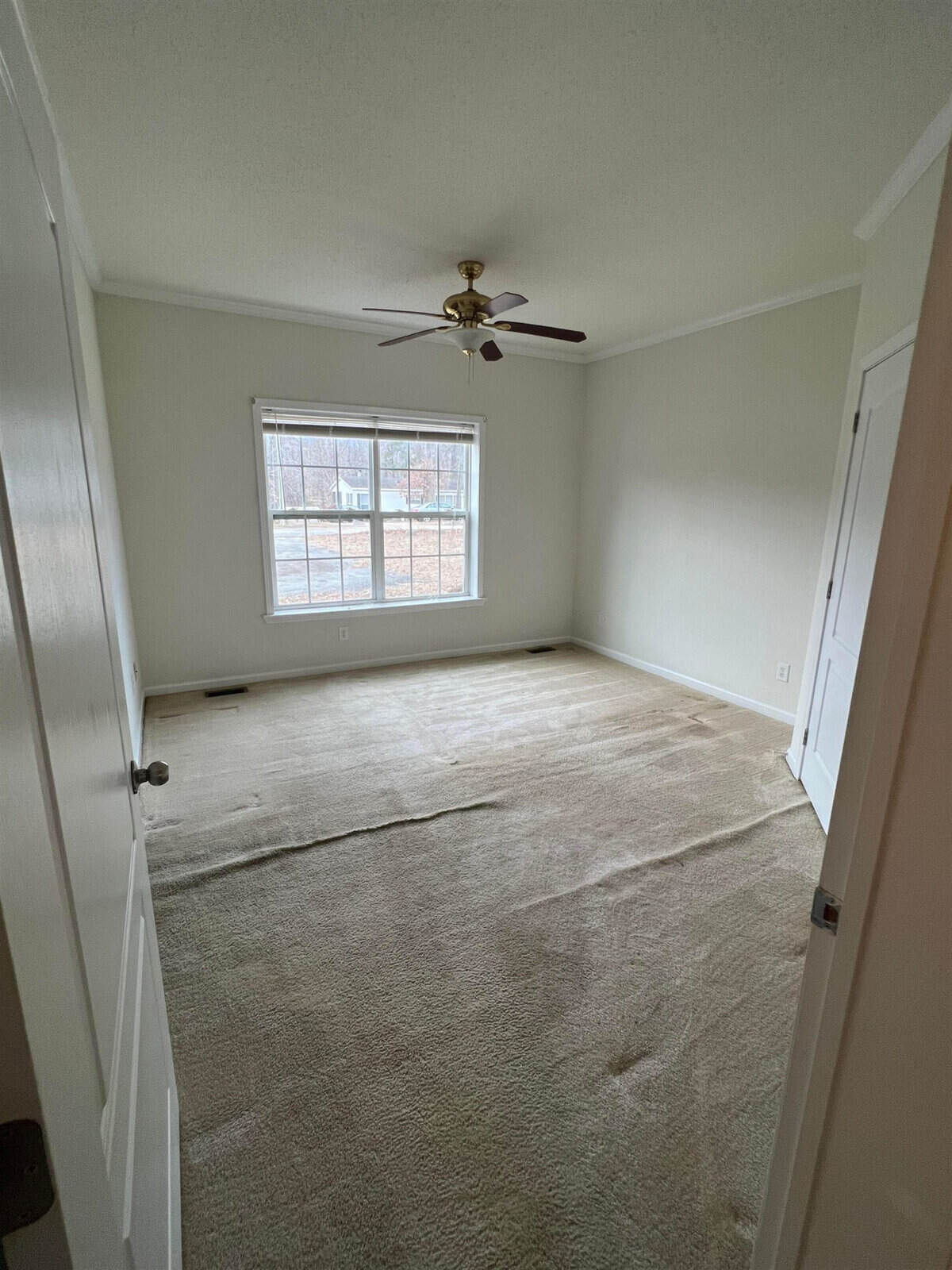 An empty room with beige carpet, white walls, and a ceiling fan with a light fixture, featuring a large window with a view of bare trees outside.
