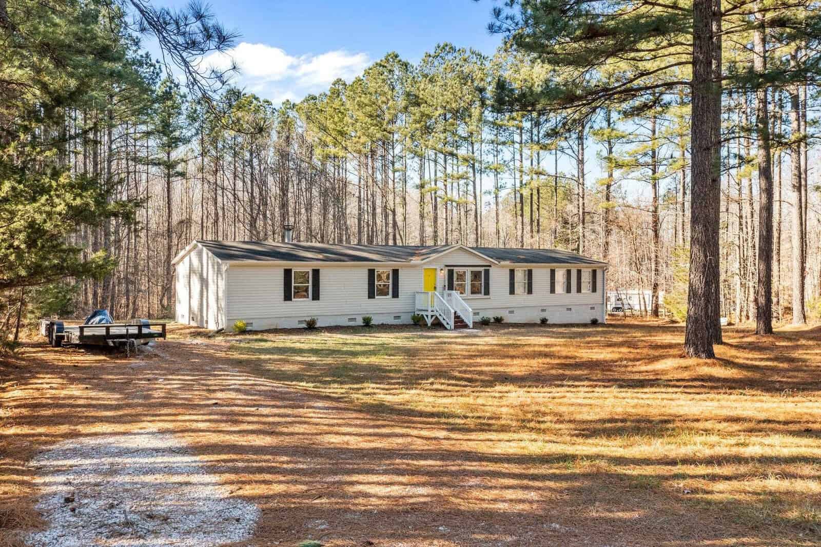 A single-story modular home at 77 Skylark Lane in Henderson NC, surrounded by tall pine trees with a clear blue sky in the background.