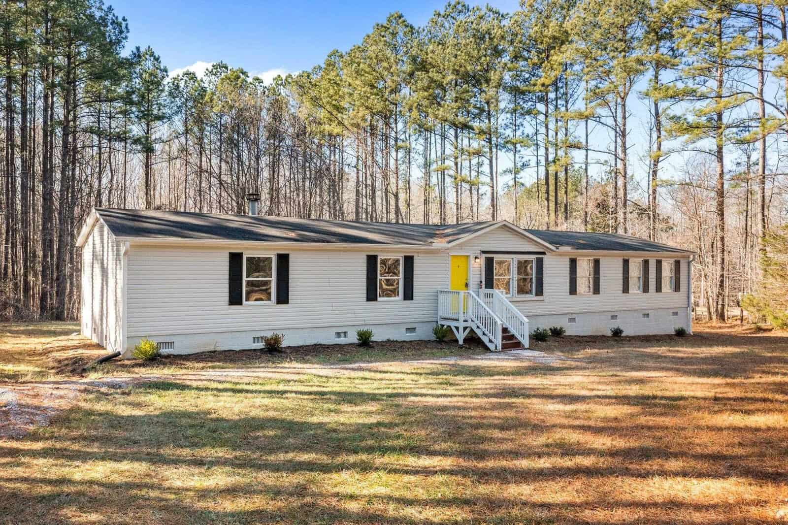 A single-story modular home with white siding and a bright yellow front door, featuring a small wooden staircase, surrounded by a grassy lawn and tall pine trees in the background.