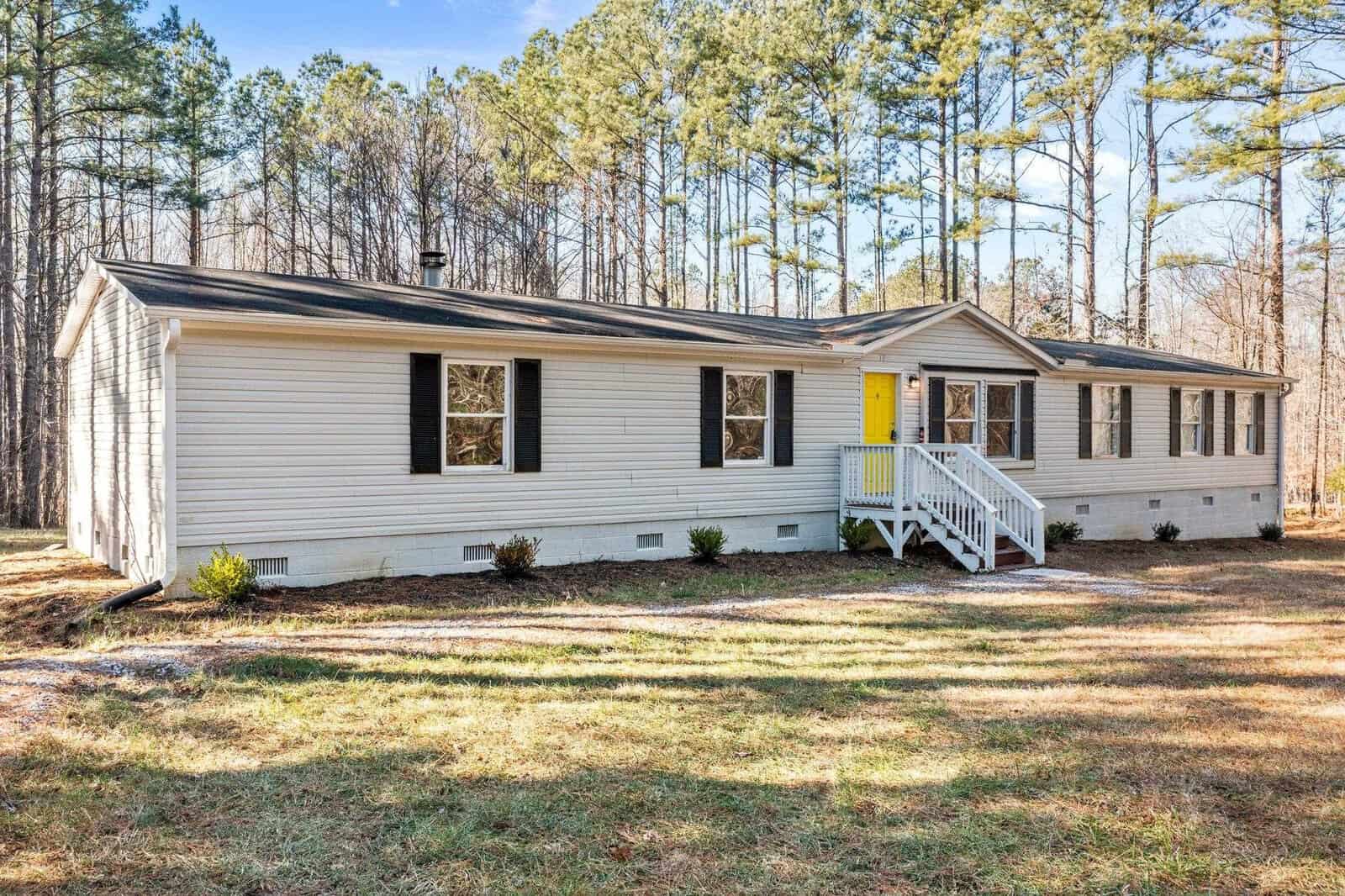 A single-story modular home with white siding and a yellow front door, featuring a small front porch with white railings, surrounded by a grassy lawn and set against a backdrop of tall trees in a sunny, clear setting.