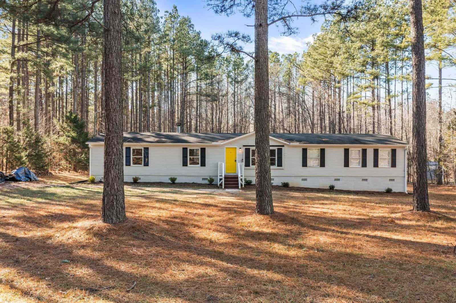 A single-story white house with a yellow front door, situated in a clearing surrounded by tall pine trees under a clear blue sky.