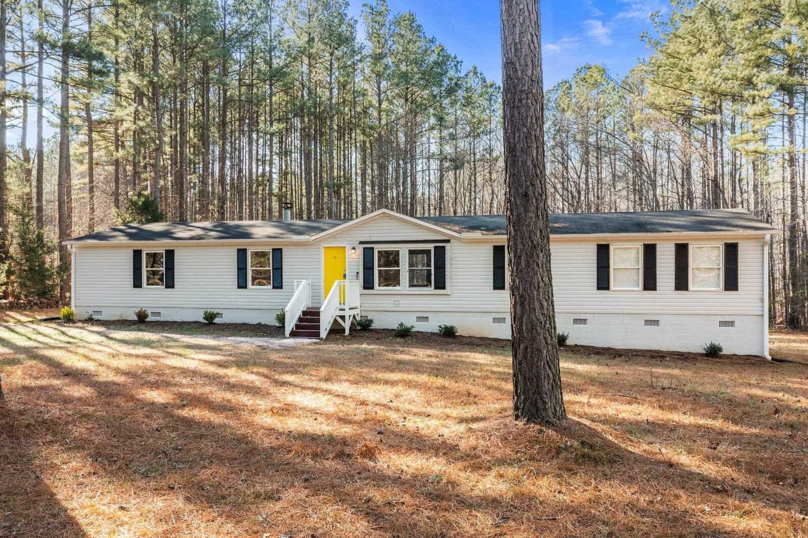 A single-story white house with a bright yellow door, surrounded by tall pine trees under a clear sky.