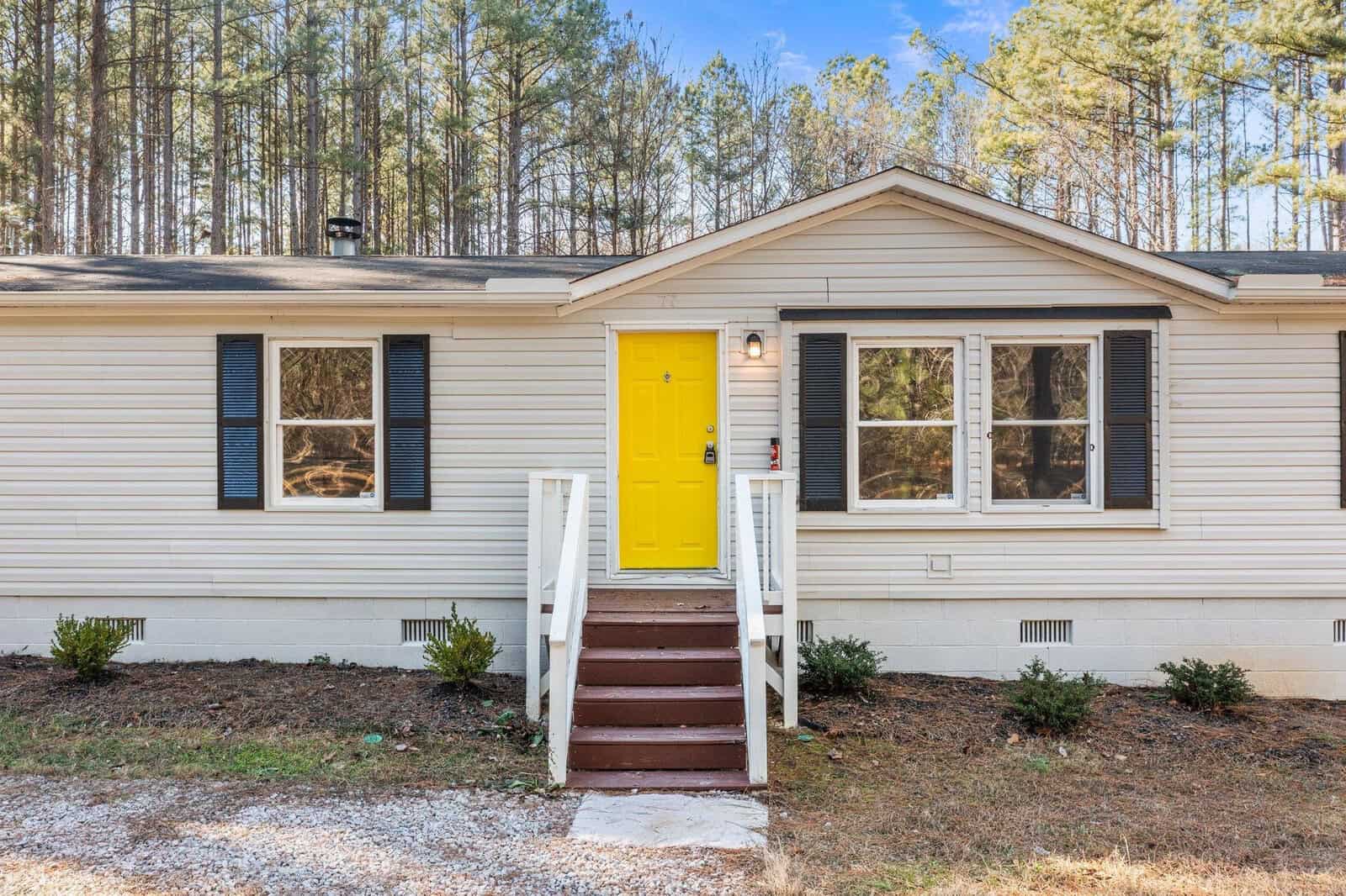 A single-story beige house with a brightly colored yellow front door, flanked by two sets of double windows and surrounded by a wooded area.
