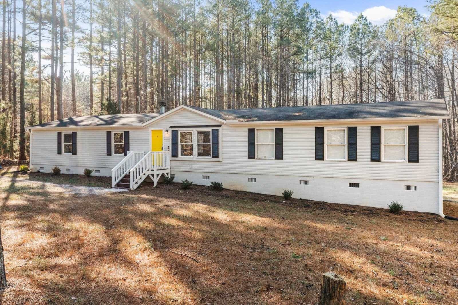A single-story modular home with white siding and a yellow front door, featuring a small wooden staircase, surrounded by a wooded area.