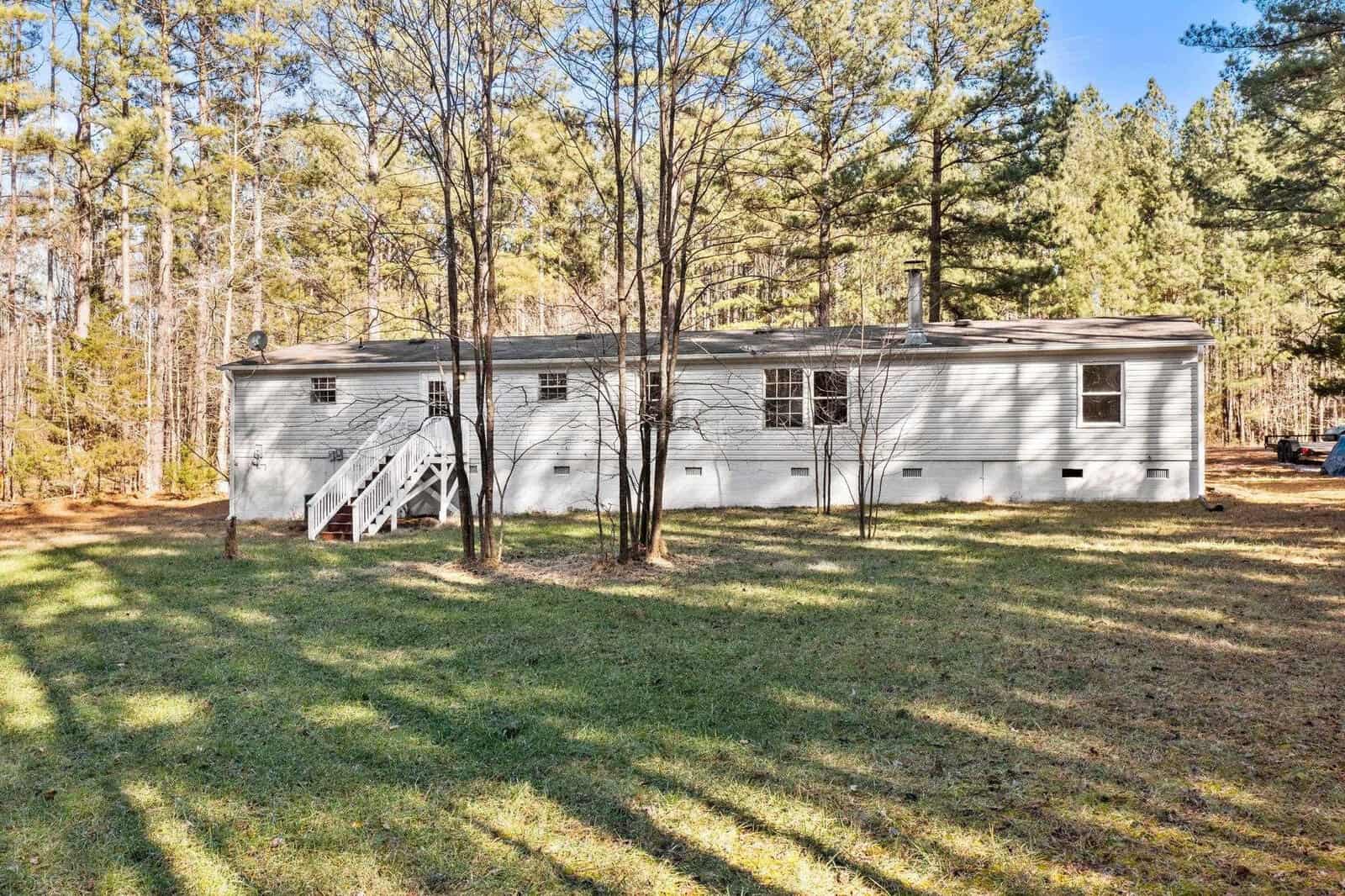 A single-story gray house with white trim situated in a clearing surrounded by tall pine trees, featuring a staircase on its side leading to a back entrance.