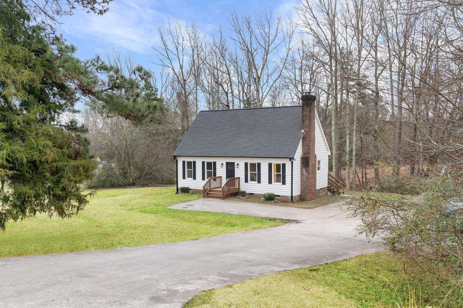 A quaint single-story house with a large chimney and white siding sits surrounded by trees on 1805 St Andrews Church Road, Henderson NC, featuring a clear front yard and a curved driveway.