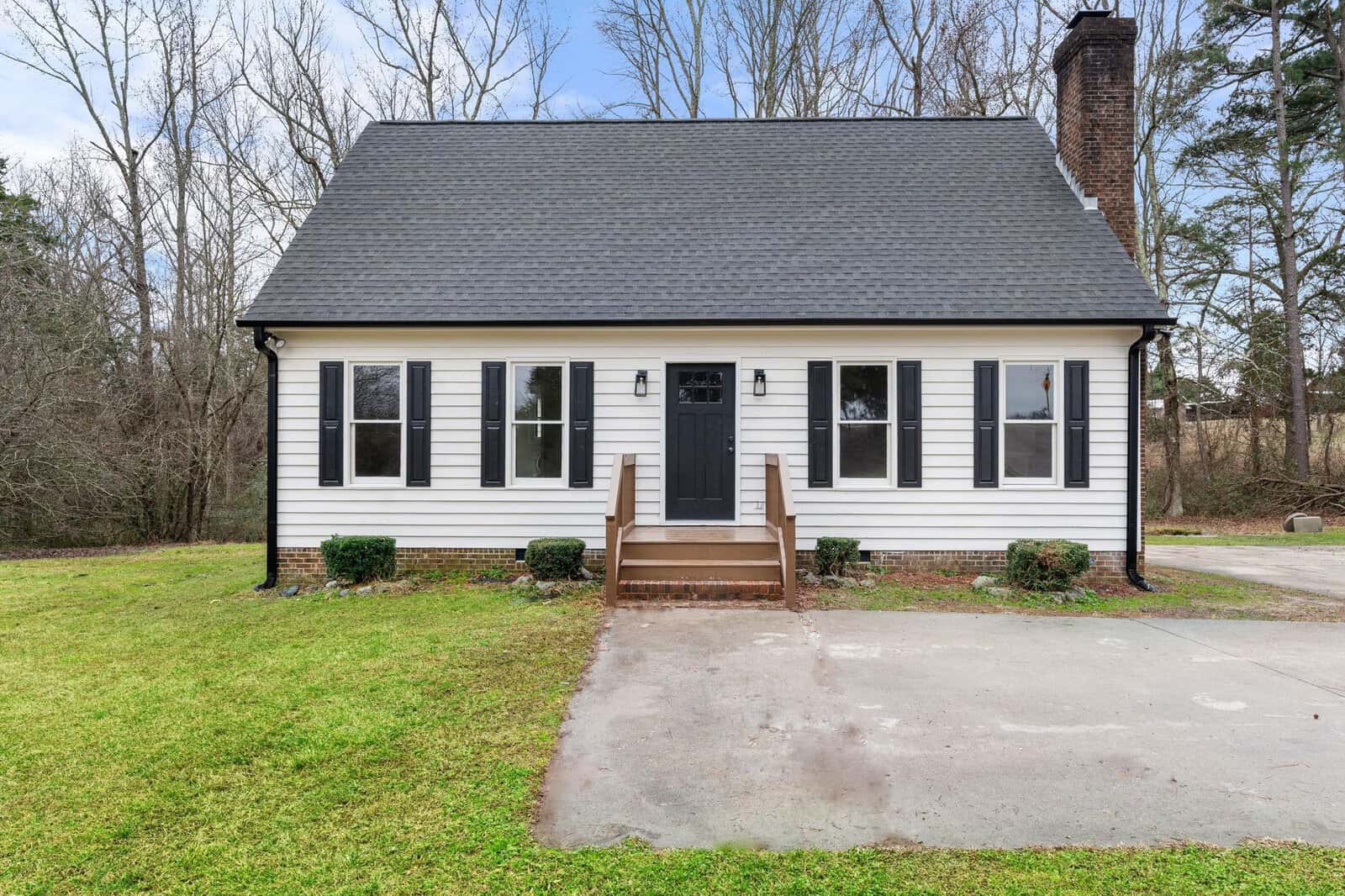 A small, two-story suburban house with white siding, black shutters, and a dark shingled roof. there's a black front door with steps leading up to it, a chimney on the left side, and a bare tree in the background. the house has a front lawn with a few low shrubs near the foundation.