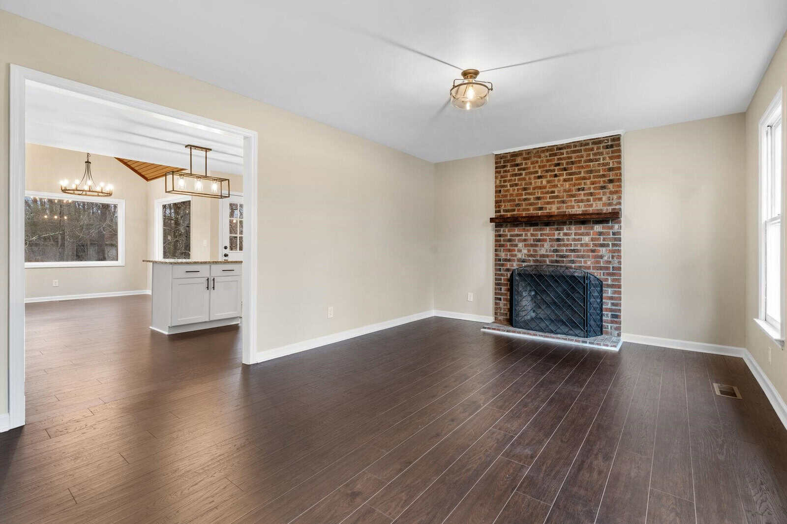A spacious, empty room with hardwood floors featuring a brick fireplace and an open doorway leading to a kitchen with white cabinetry and hanging lights.
