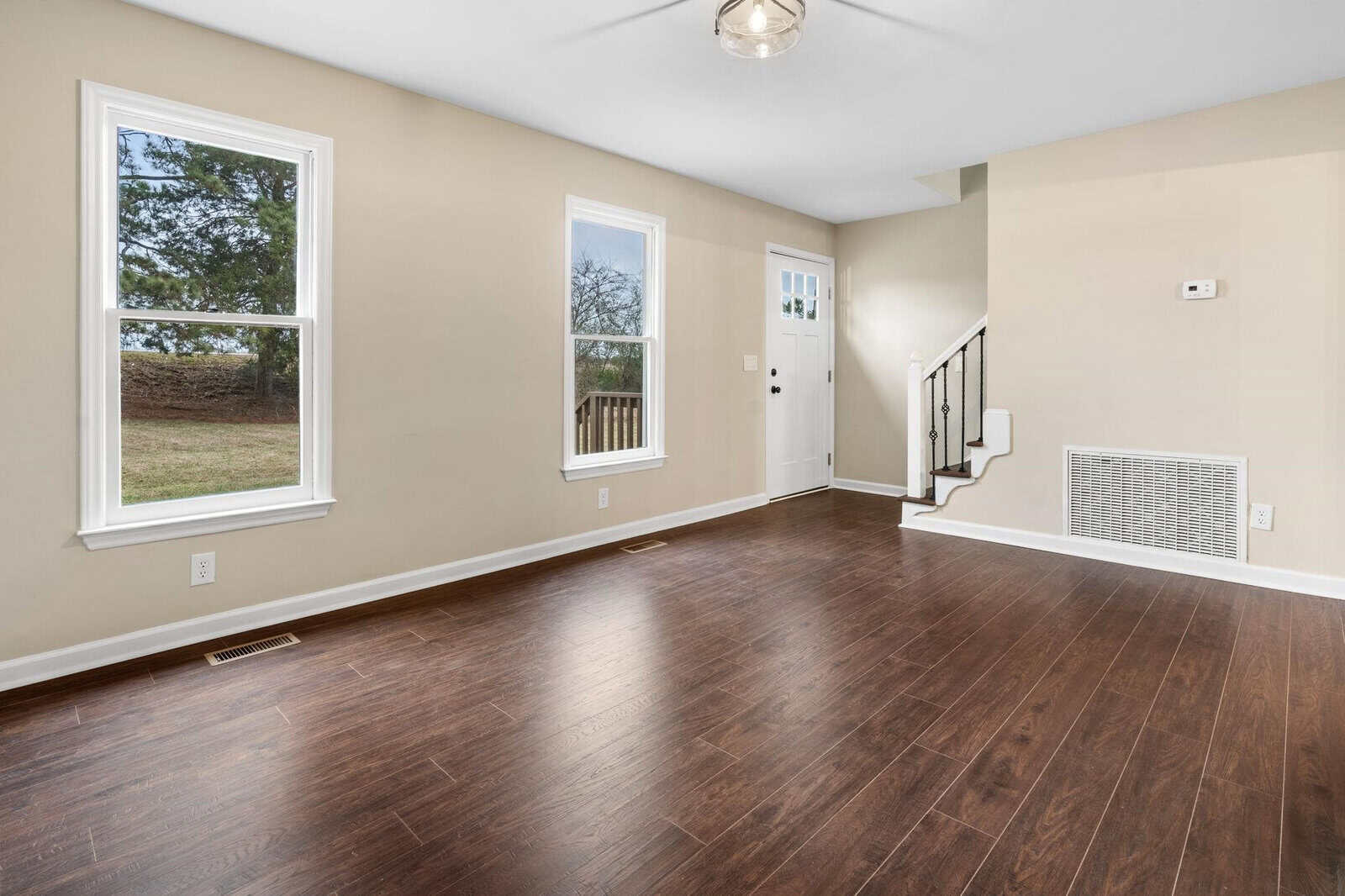 Empty room with hardwood floors, a white door with sidelights, three windows, and a staircase on the right.