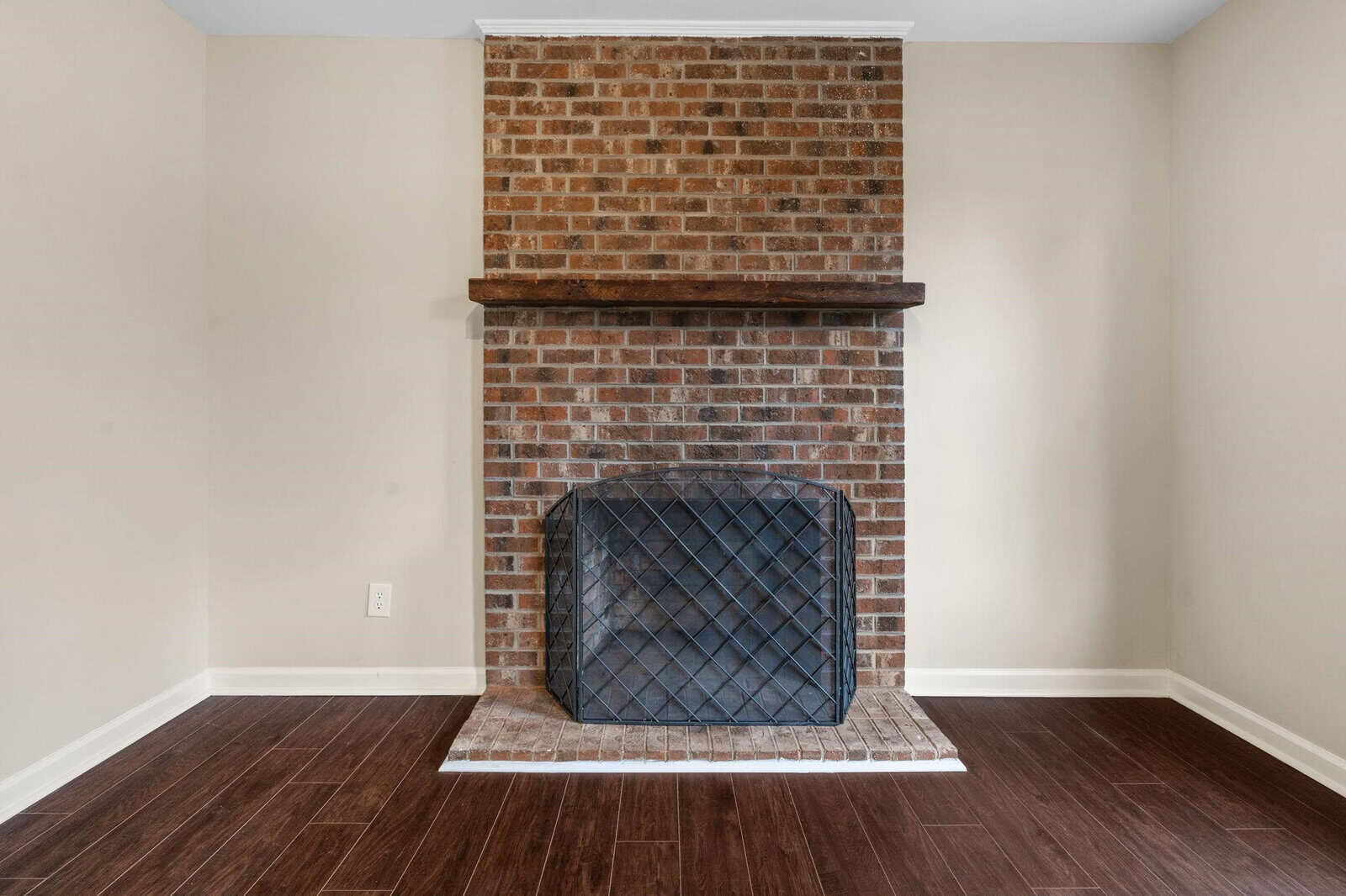 This image shows an interior room corner featuring a brick fireplace with a dark wooden mantel, a protective mesh screen, and a white hearth, set against walls painted in a neutral color and a dark hardwood floor.