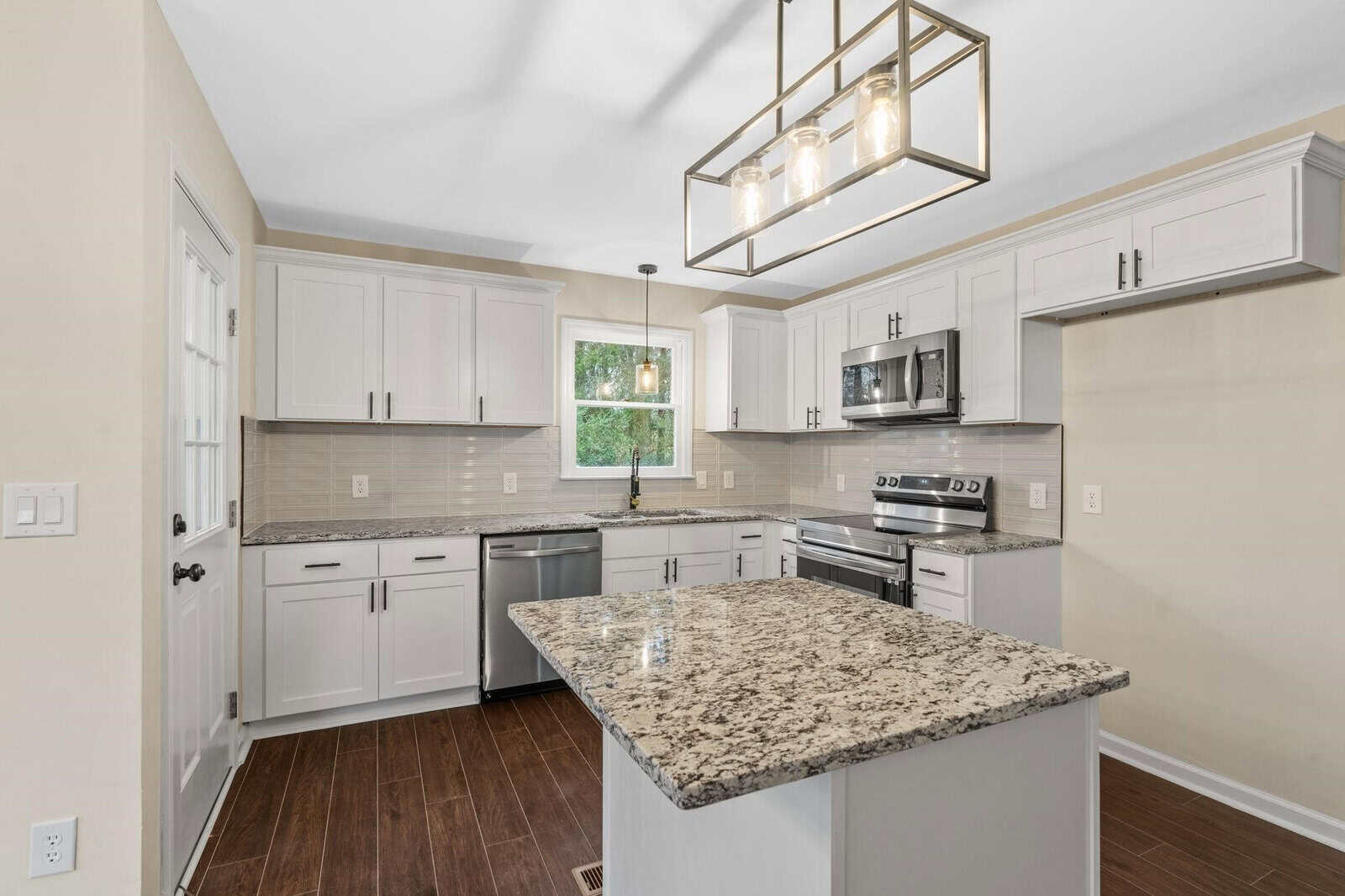A modern kitchen with white cabinetry, stainless steel appliances, granite countertops, and a central island under a hanging rectangular light fixture.