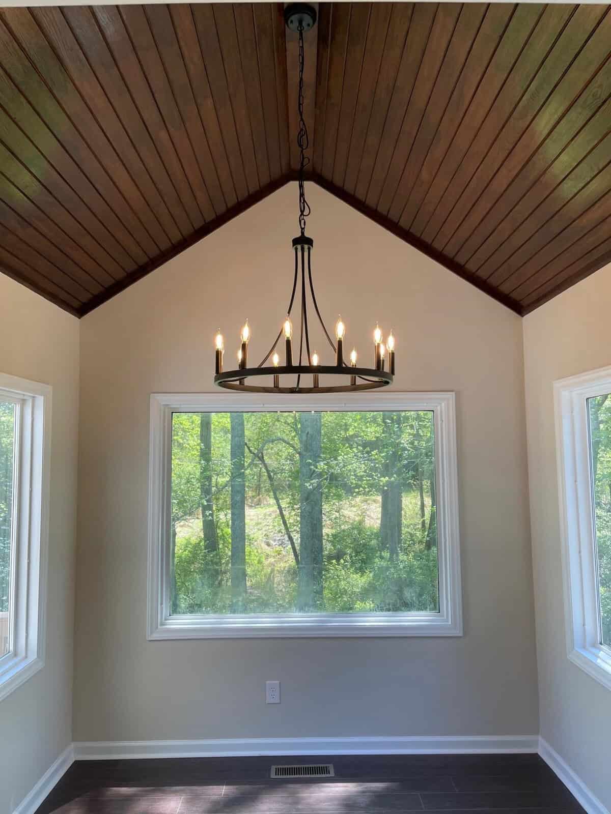 A spacious room with dark hardwood floors and white walls, featuring a large window with a view of greenery, under a vaulted ceiling with a wooden finish and a hanging chandelier with lit candles.