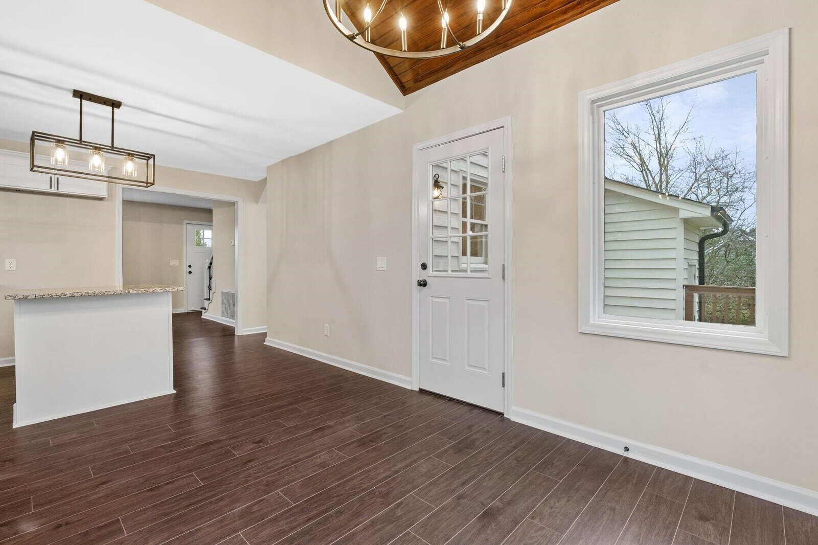 An empty room featuring hardwood flooring, white walls, a large window, and a white front door, with a view into an adjoining room and a modern chandelier overhead.
