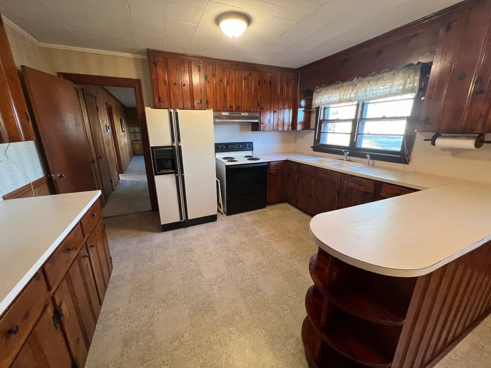 A spacious kitchen with wooden cabinetry, white countertops, a refrigerator, stove, and a window with curtains above the sink.