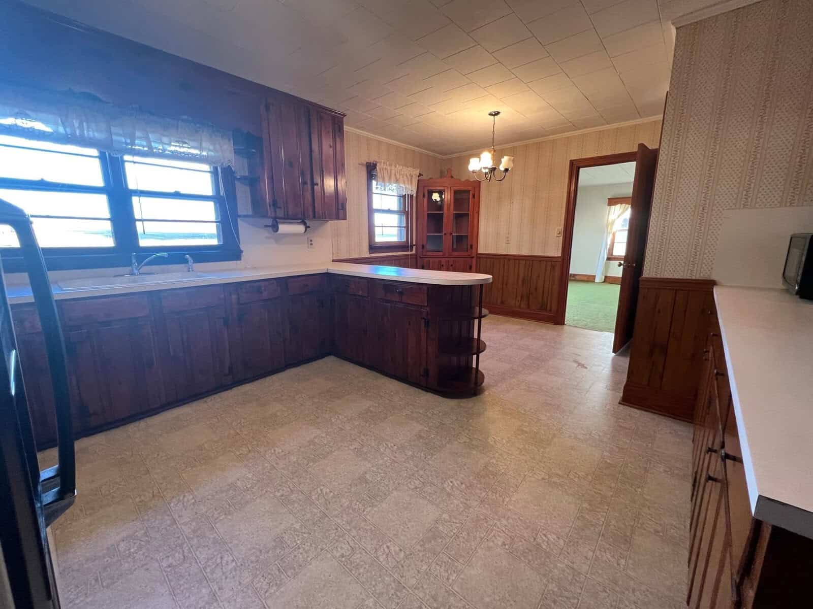 An empty kitchen with wooden cabinets, a white countertop, and linoleum flooring, featuring a breakfast bar area and green carpet in the adjacent room.