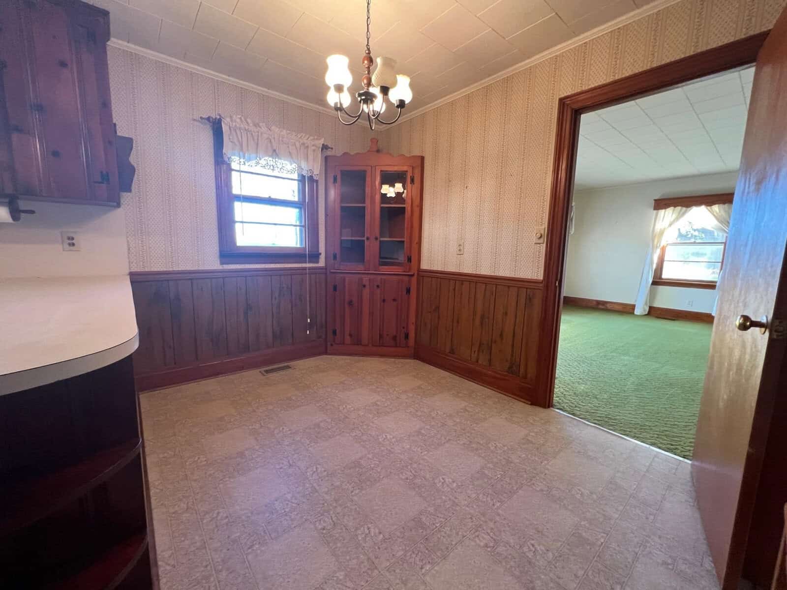 An empty room featuring a wooden wainscot, a built-in glass-fronted cabinet, and a chandelier, with an adjoining room visible through an open doorway.