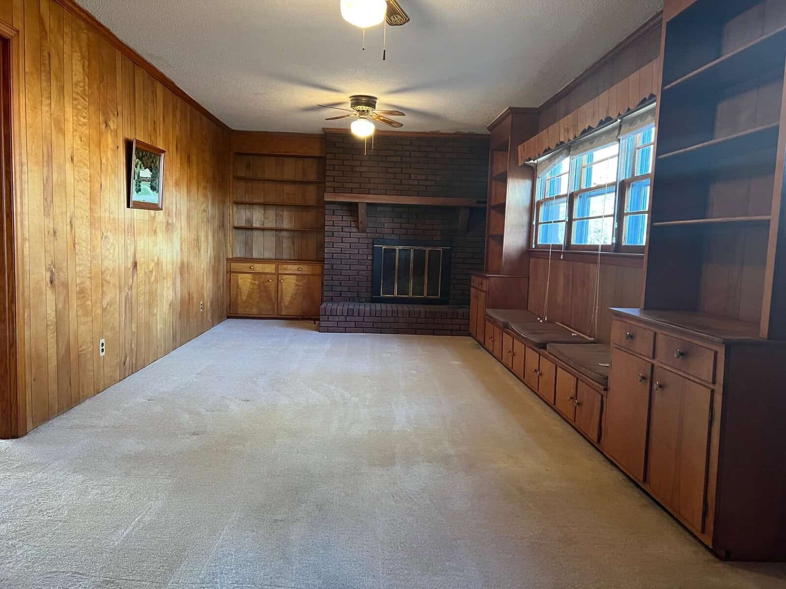 An empty room with wood paneling, a brick fireplace, carpeted flooring, built-in shelves, and a window with blinds.