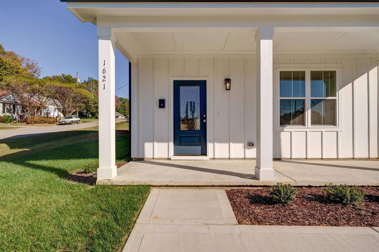 A modern front porch of a house with the number 1621 displayed, featuring a dark-colored front door and two windows beside it, set against white exterior walls with a clear blue sky above and a neatly manicured lawn in front.