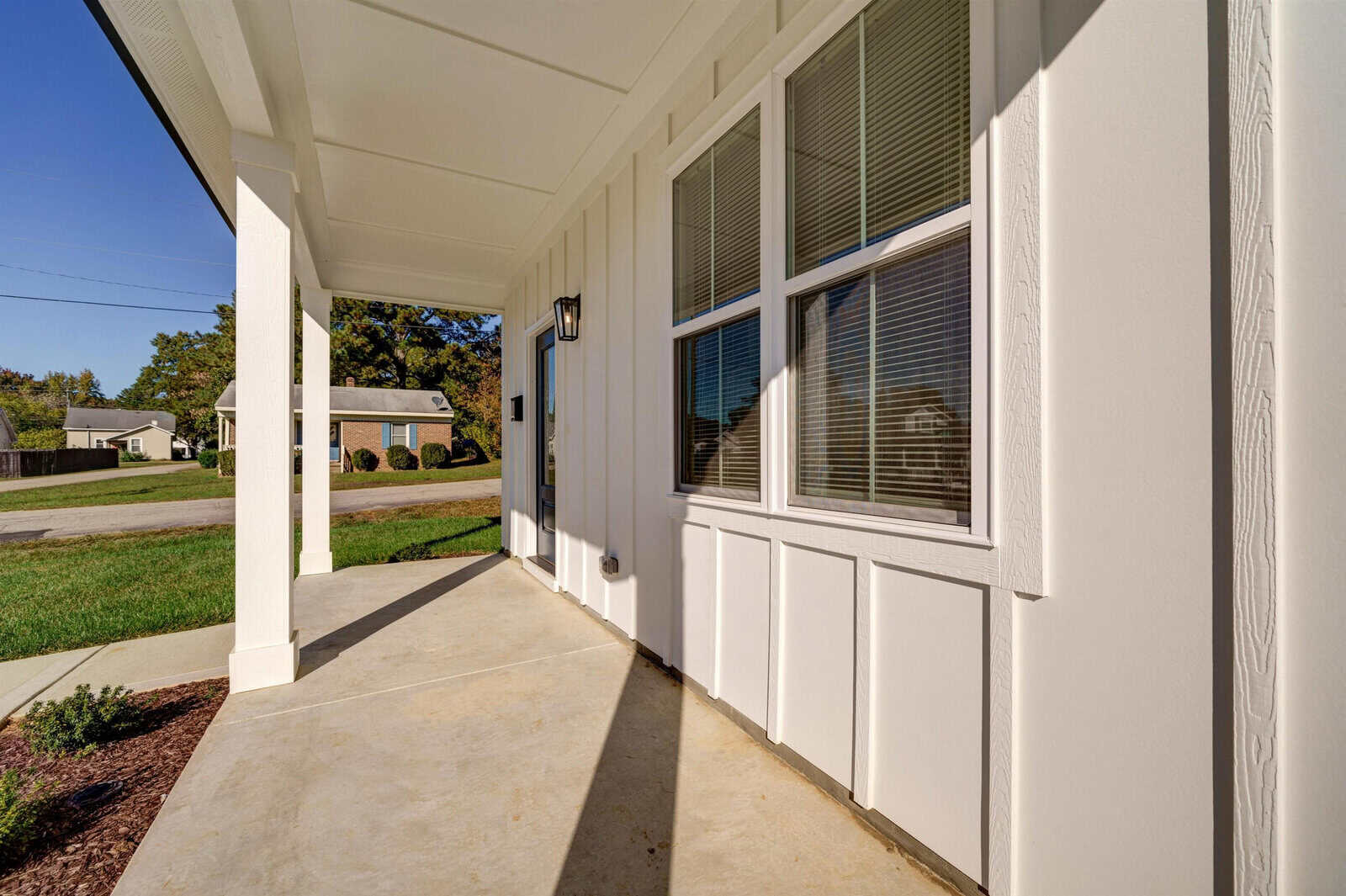 A sunny view of a residential home's entrance, featuring a concrete walkway, white exterior walls, and large windows with blinds.