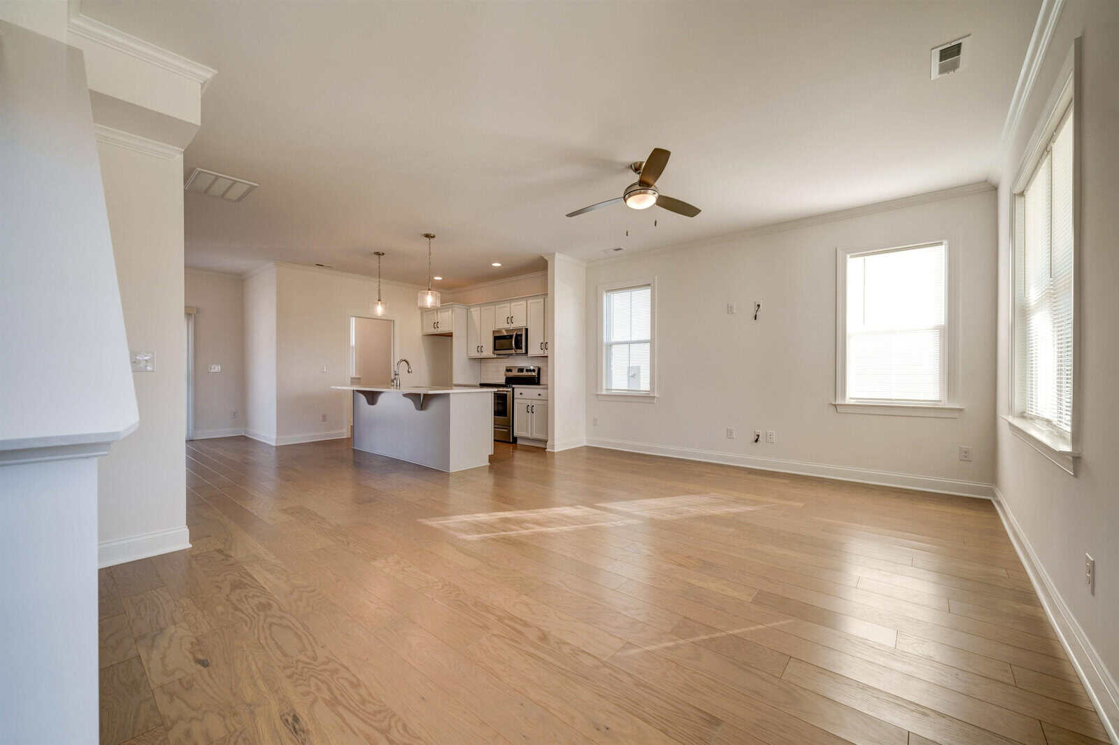 A spacious, empty room with hardwood floors, white walls, and a kitchen area in the background. the room is well-lit with natural light from multiple windows and is equipped with a ceiling fan.