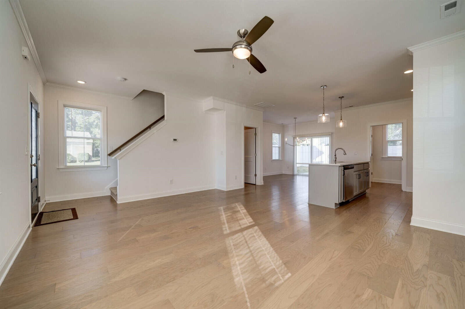 Spacious, well-lit room with wooden flooring featuring a staircase, ceiling fan, and kitchenette with modern fixtures.