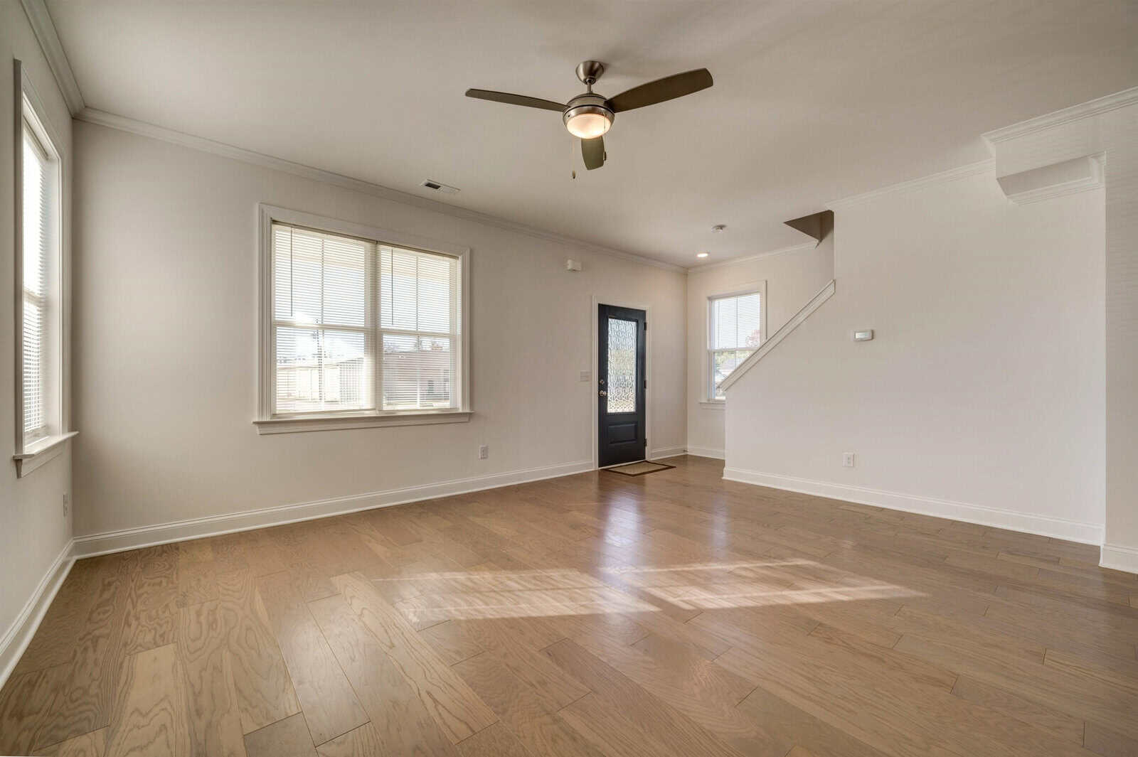 An empty room with hardwood floors, a ceiling fan, natural light coming through windows, and a staircase on the right-hand side leading to another level.